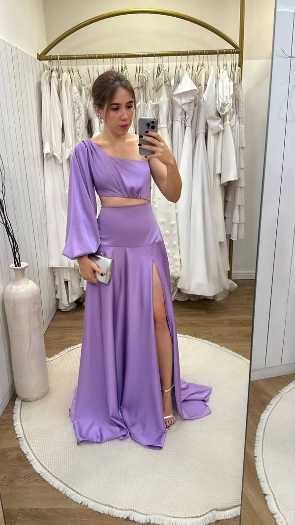 Vestido longo lilás em tecido acetinado, modelo um ombro com manga longa, recorte na cintura e fenda alta na saia.