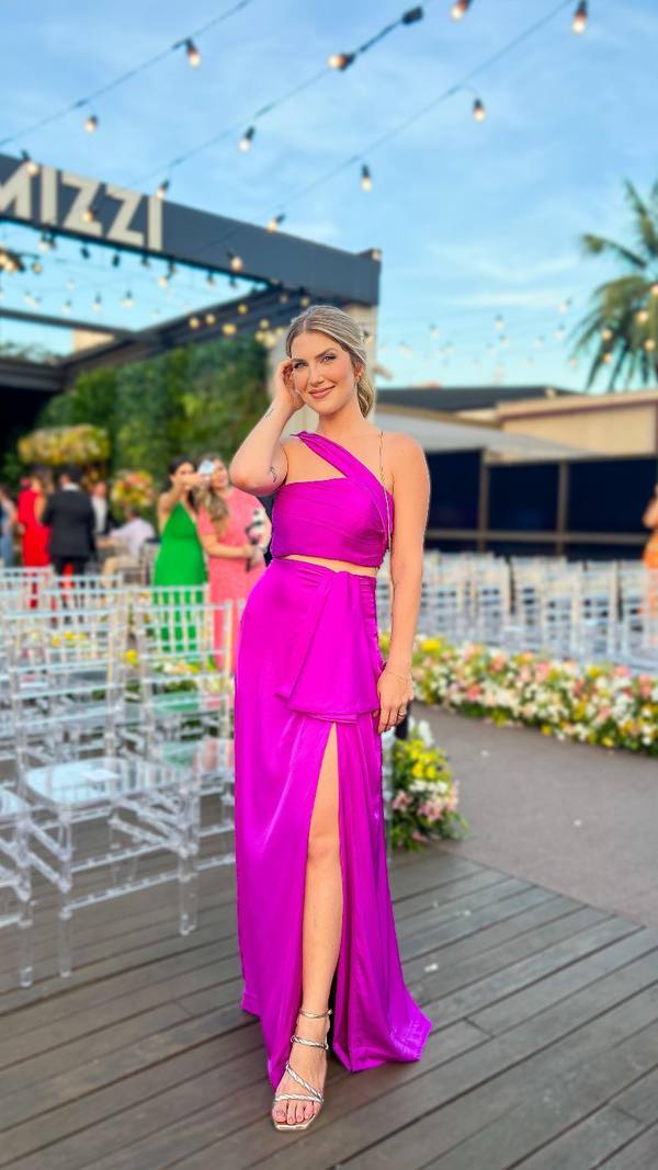 Vestido longo fúcsia de cetim, com decote de um ombro só, fenda pronunciada na saia e caimento fluido.