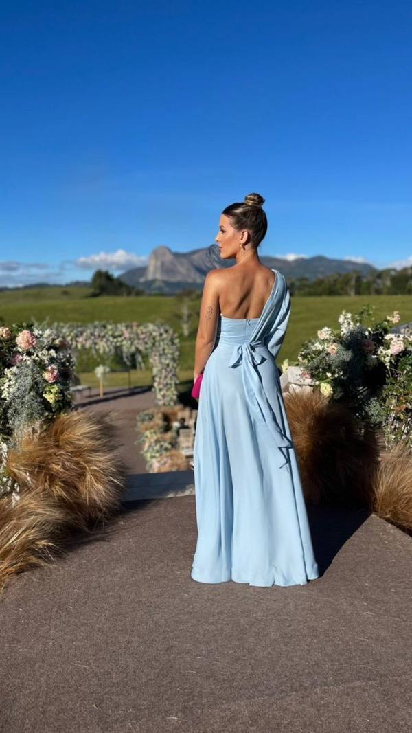 Vestido longo azul claro de um ombro só, com caimento fluido e detalhe de laço nas costas.