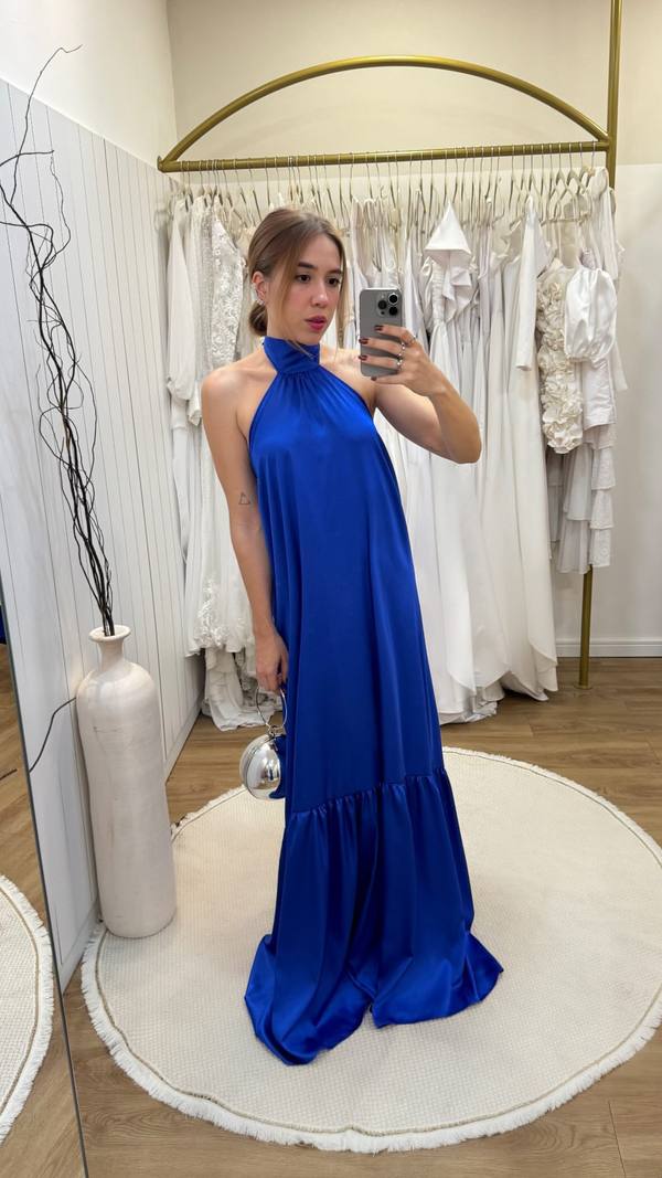 Vestido longo azul de cetim, com decote frente única, modelagem fluida e detalhe de babado na barra.