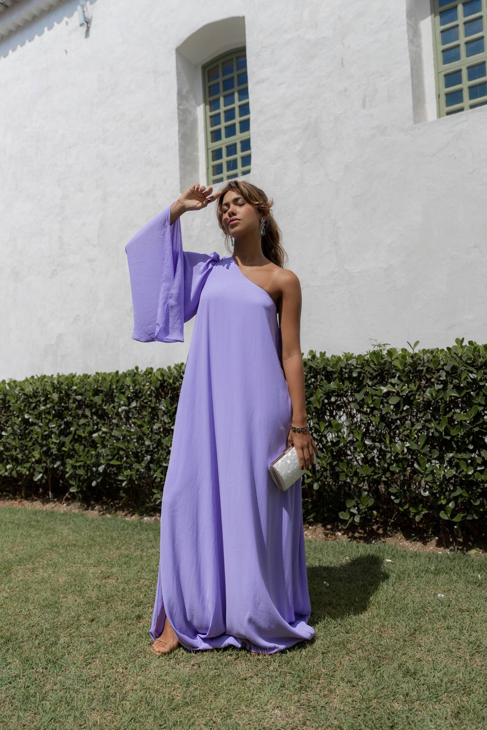 Vestido longo lilás de caimento fluido, modelagem um ombro com manga ampla e caimento leve.