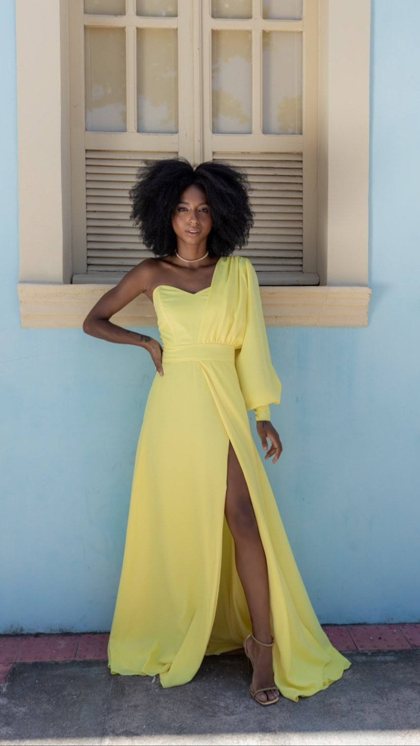 Vestido longo amarelo de um ombro só, com manga longa bufante, cintura marcada e fenda profunda na saia fluida.