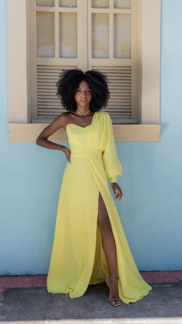 Vestido longo amarelo de um ombro só, com manga longa bufante, cintura marcada e fenda profunda na saia fluida.