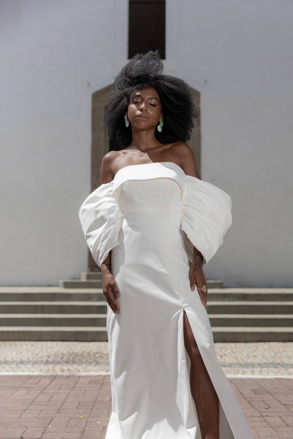 Vestido longo branco tomara que caia, com mangas bufantes caídas, fenda frontal e caimento reto.