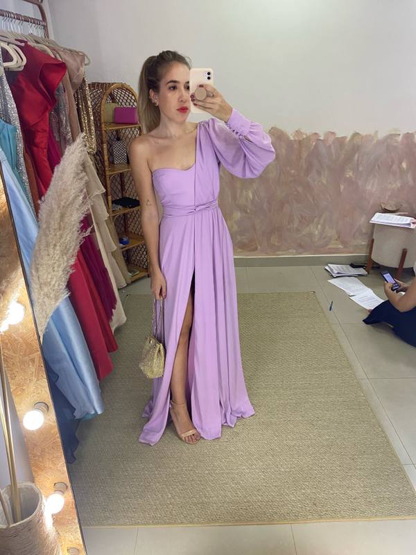 Vestido longo lilás de caimento fluido, modelo um ombro com manga longa, cintura marcada e fenda na saia.