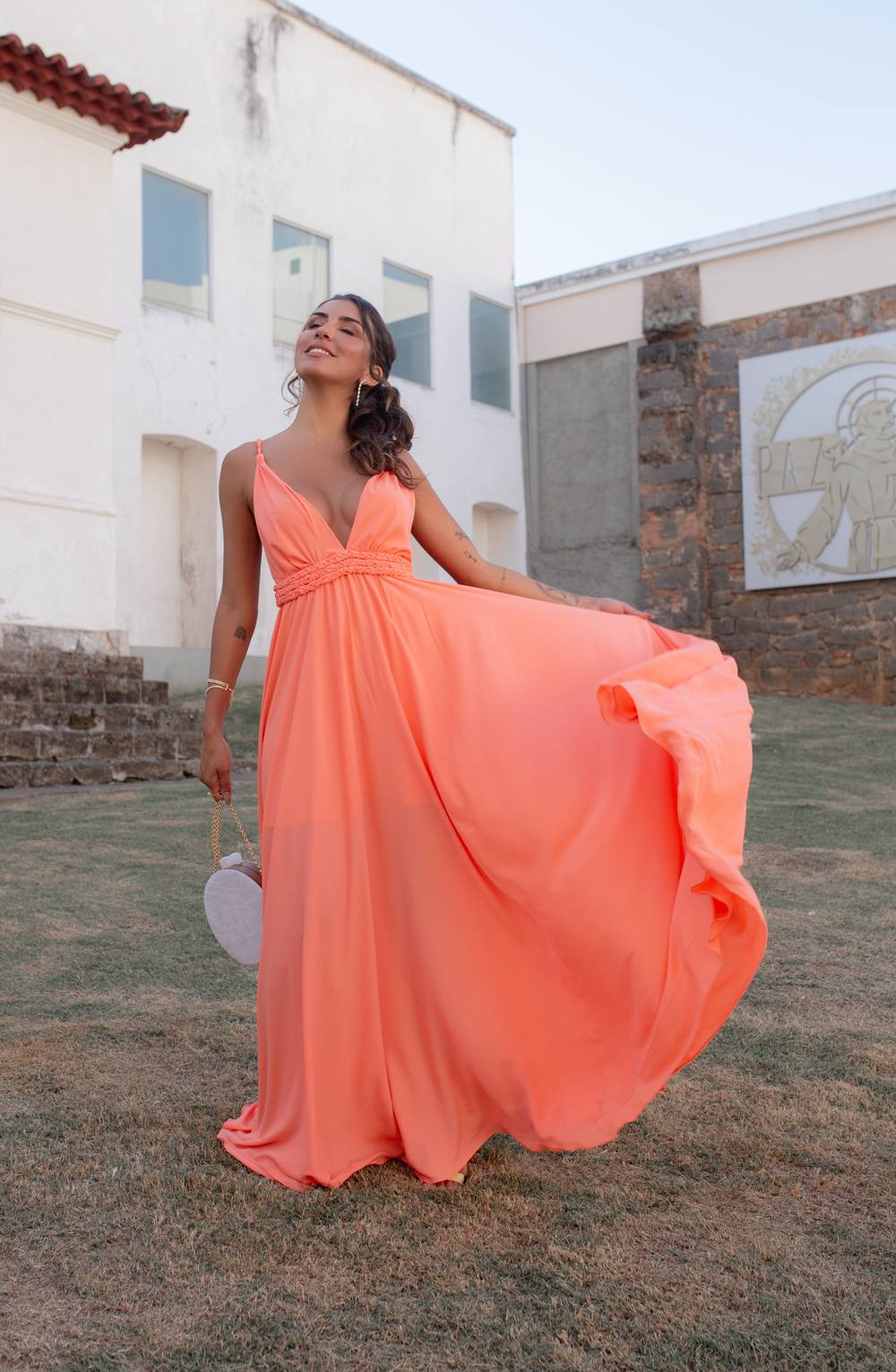 Vestido longo coral de alças finas e decote V, em tecido fluido com detalhe trançado abaixo do busto e saia ampla.