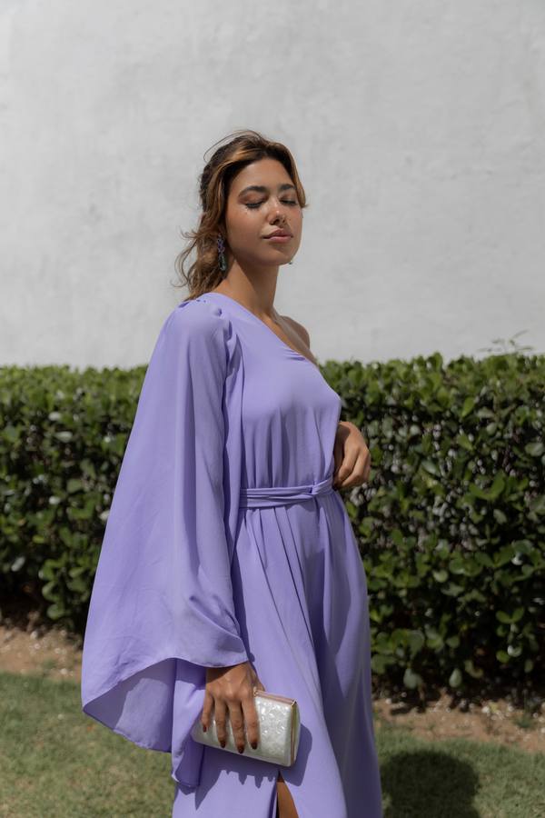 Vestido longo lilás de um ombro só, caimento fluido com manga ampla, faixa na cintura e fenda na saia.