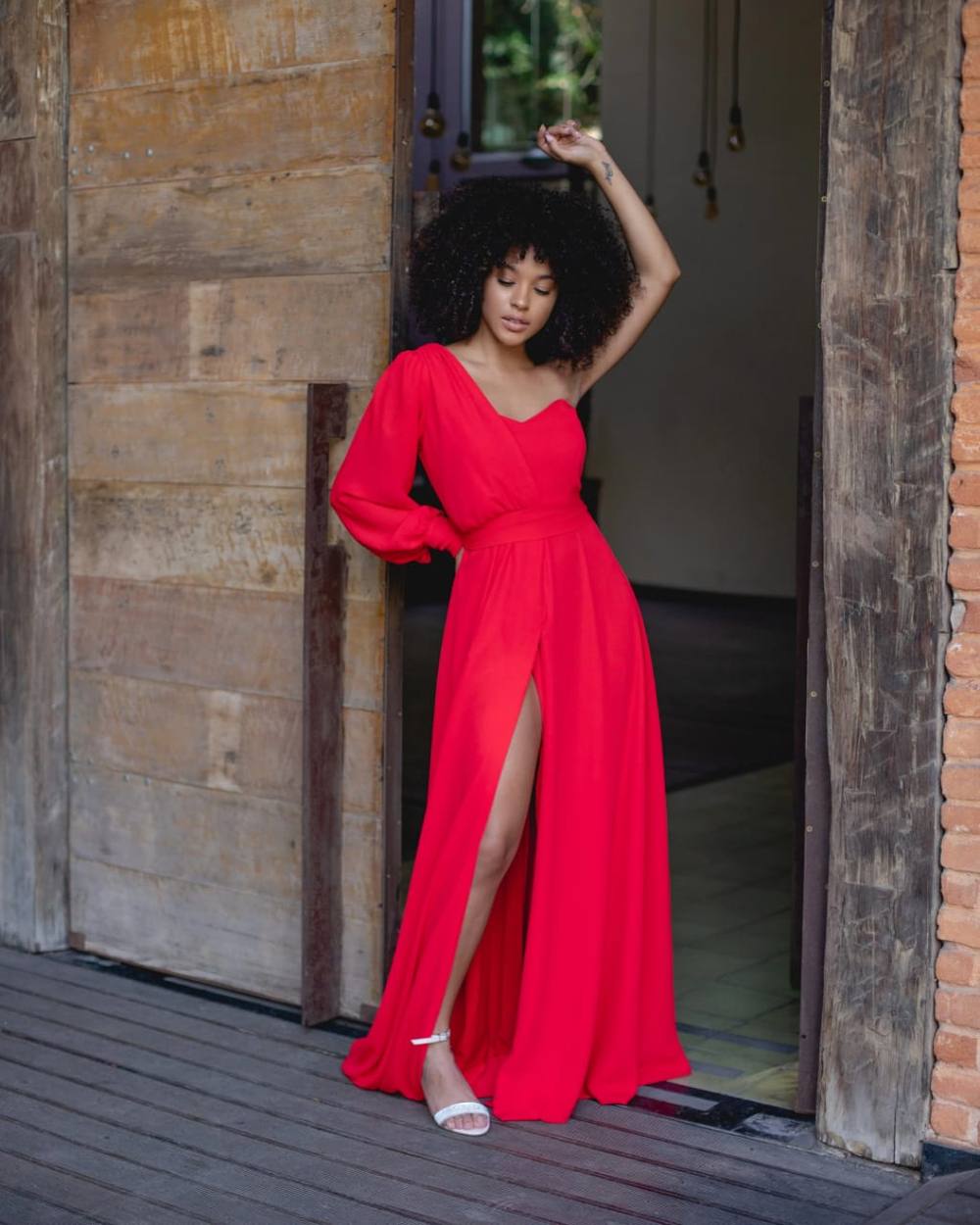 Vestido longo vermelho em tecido fluido, decote um ombro com manga longa, cintura marcada e fenda frontal na saia.