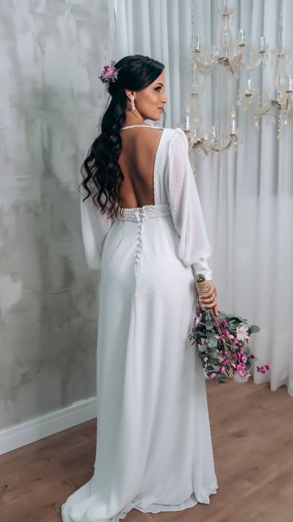 Vestido longo branco com mangas longas, costas abertas, saia fluida em tecido leve e botões na parte traseira.