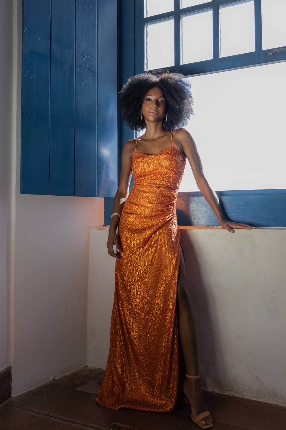 Vestido longo laranja de paetês, com alças finas, modelagem ajustada e fenda lateral na saia.