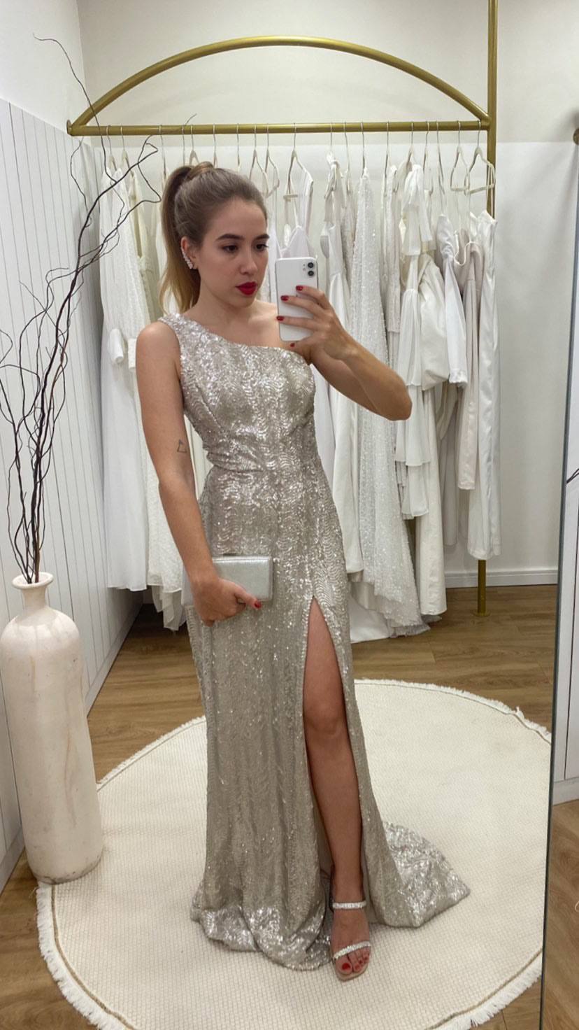 Vestido longo prata de paetês, com decote de um ombro só, fenda frontal e caimento reto.