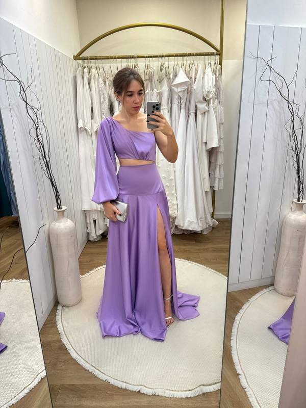 Vestido longo lilás em tecido acetinado, modelo um ombro com manga longa, recorte na cintura e fenda profunda na saia.