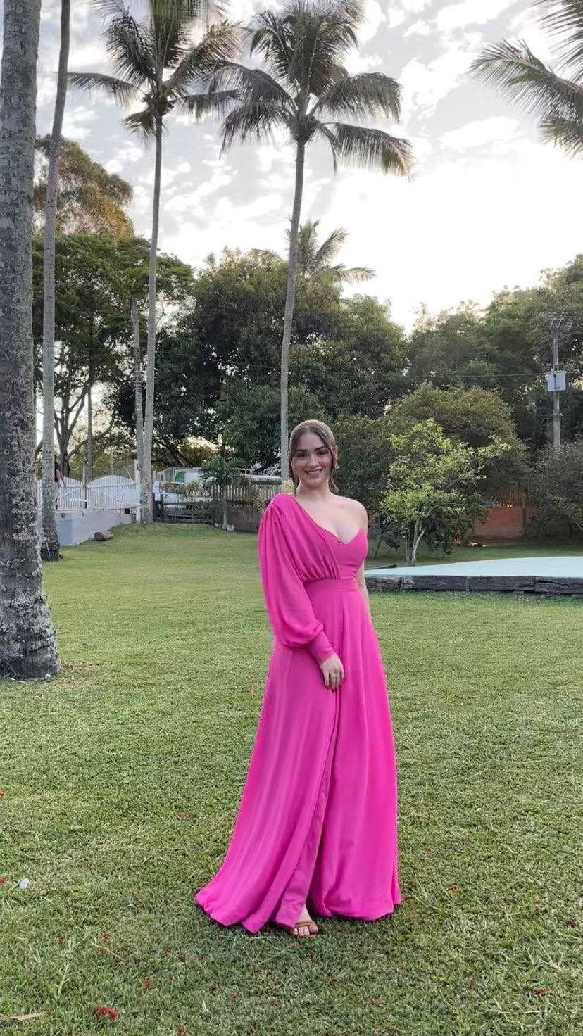 Vestido longo fúcsia fluido, com modelo um ombro de manga longa, cintura marcada e fenda na saia.