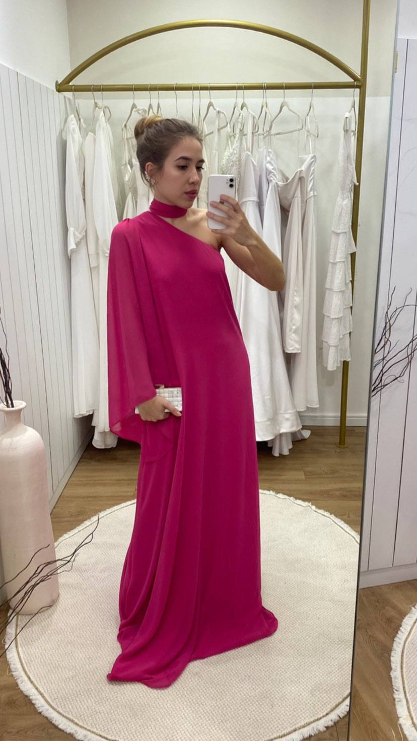 Vestido longo fúcsia de caimento fluido, modelo um ombro com faixa no pescoço e manga ampla esvoaçante.