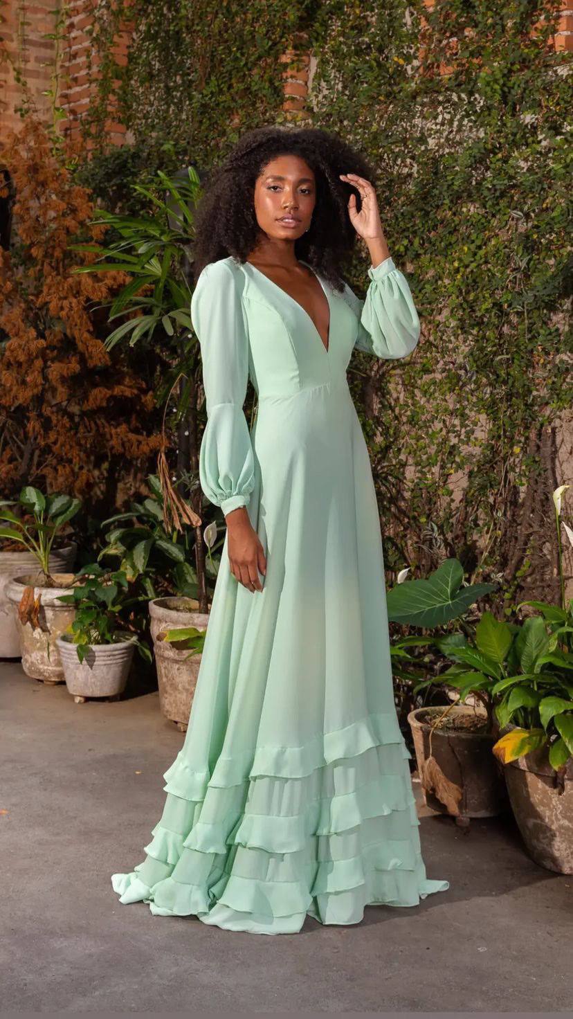 Vestido longo verde claro em tecido fluido, com mangas longas, decote em V e detalhe de babados na barra da saia.