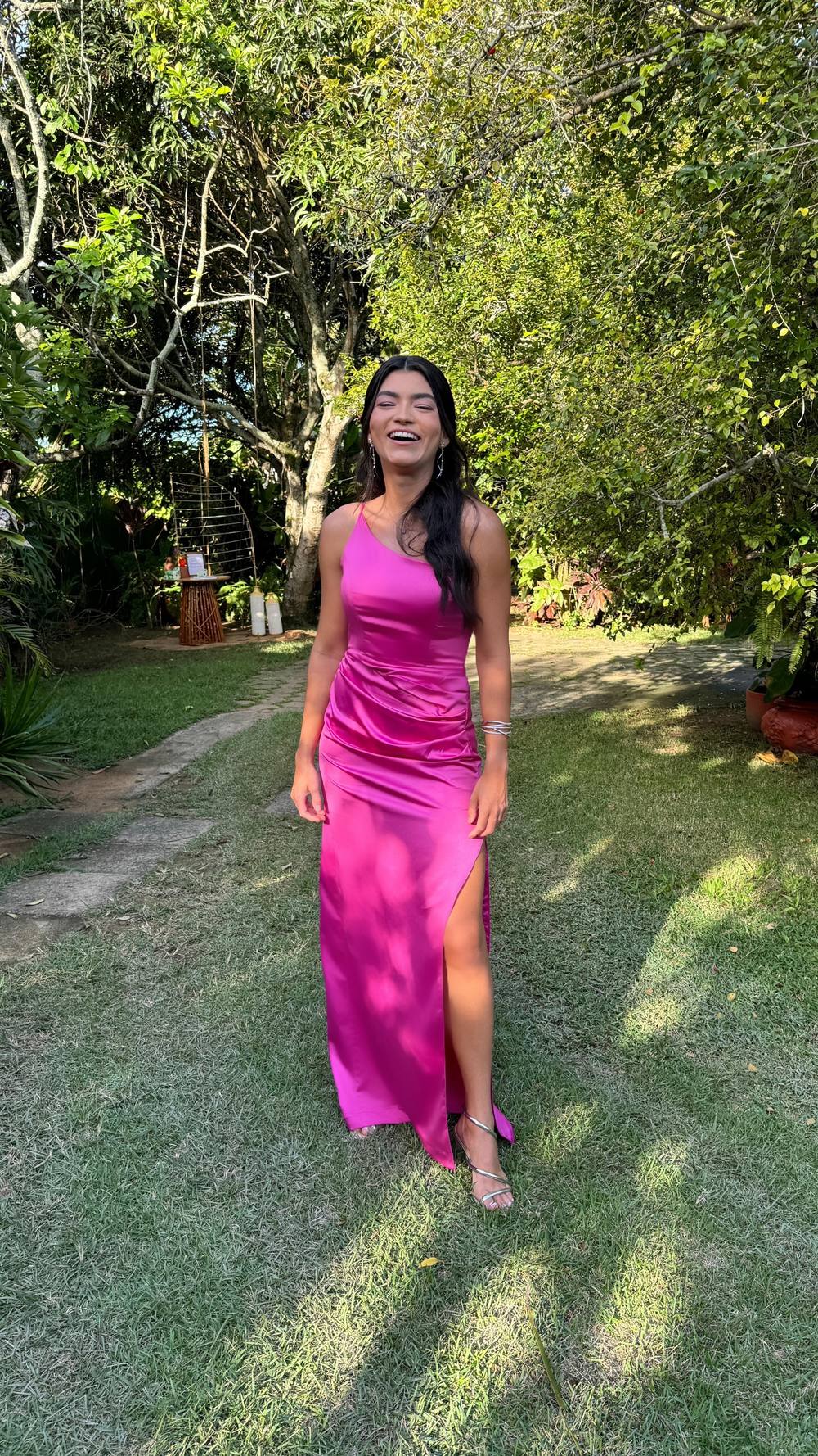 Vestido longo fúcsia em cetim, decote um ombro, caimento reto com drapeado e fenda lateral.